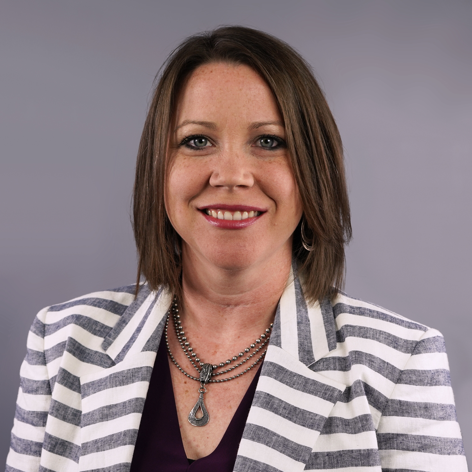 Formal headshot of a white woman wearing a striped blazer and several necklaces. She is smiling at the camera.}}