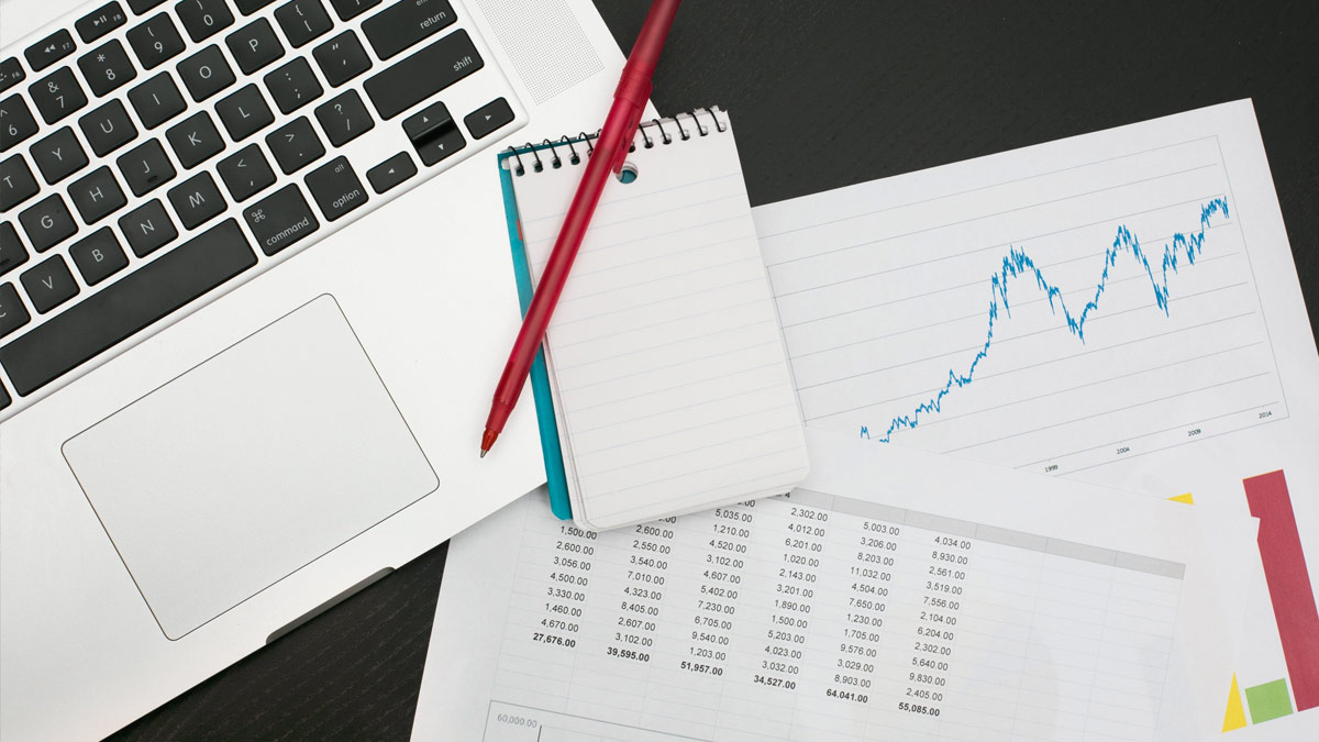 Top view of laptop with graphs printed on paper and pens sitting on a desk