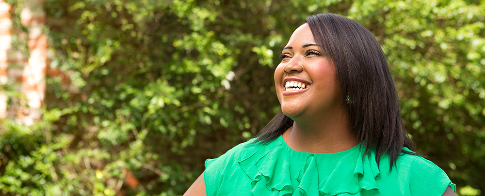 Black woman wearing green ruffle shirt standing in front of greenery smiling