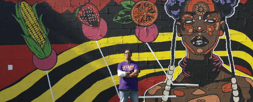 一名男子站在外面，墙上挂着一幅充满活力的非洲壁画，墙上有玉米穗、草莓和桔子片。 