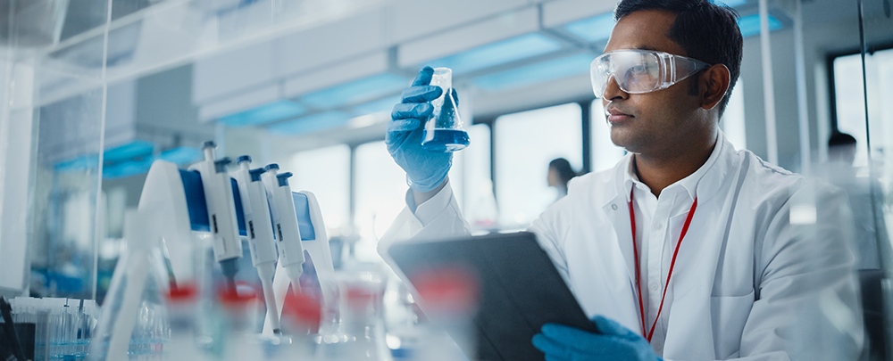 Scientist wearing white lab coat, clear safety glasses, blue gloves, raises a beaker of blue liquid in lab