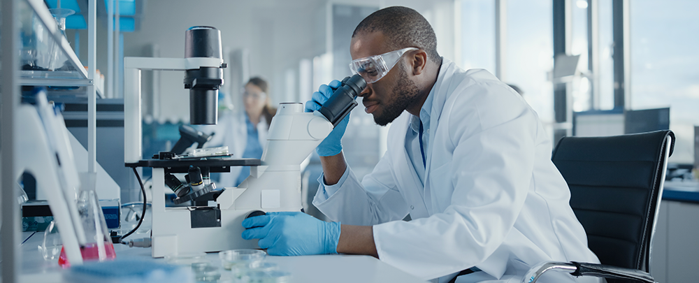 black scientist wearing lab coat, blue rubber gloves, and goggles, looking into microscope in lab
