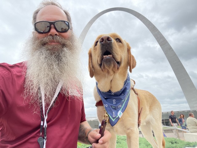 Man with a white beard wearing sunglasses, kneeling next to a large dog in a blue bandana, in front of The Gateway Arch in St. Louis