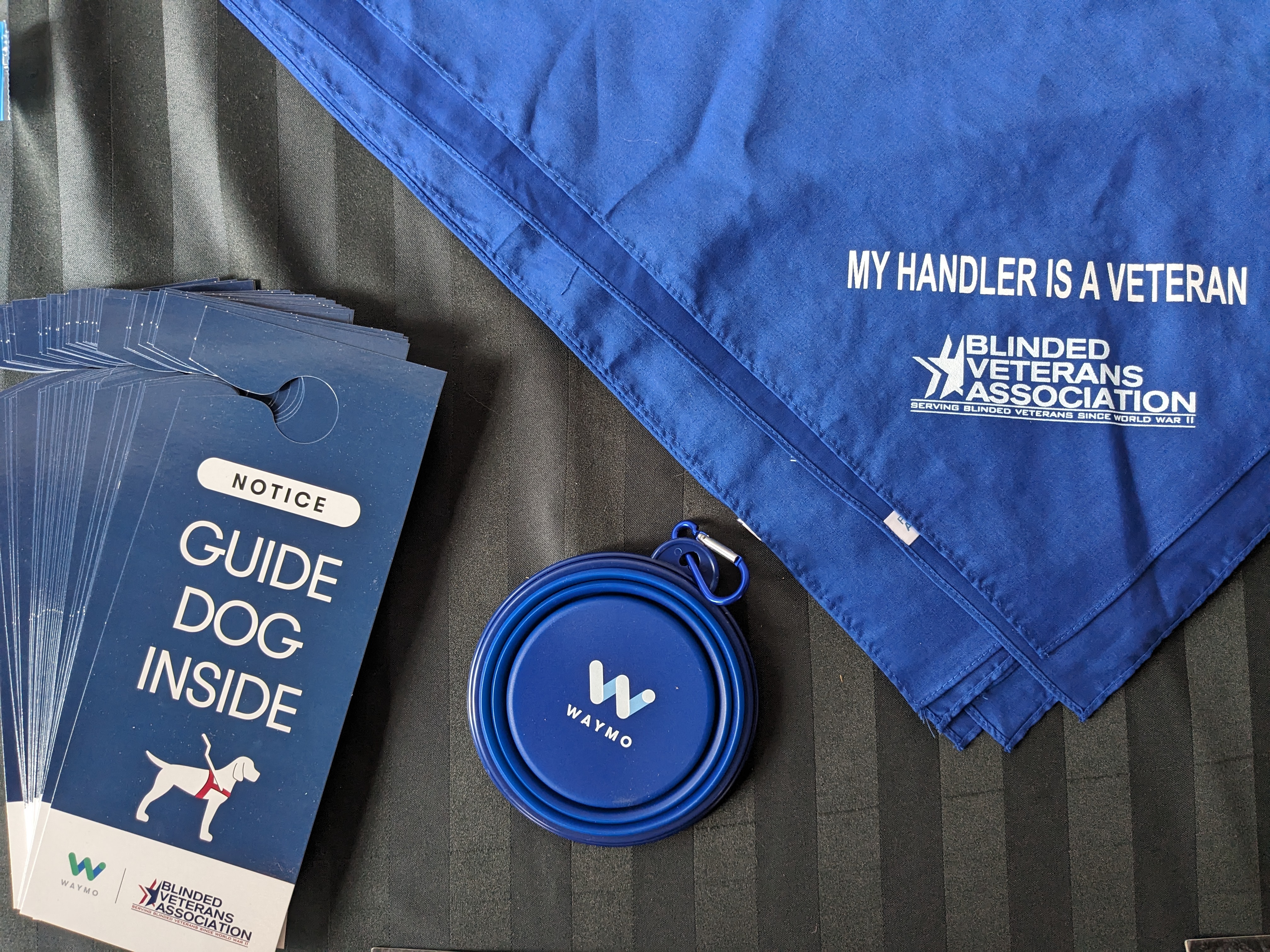 "Guide Dog Inside" Door hanger, Waymo branded collapsible water bowl, and BVA branded dog bandana laid out on a table