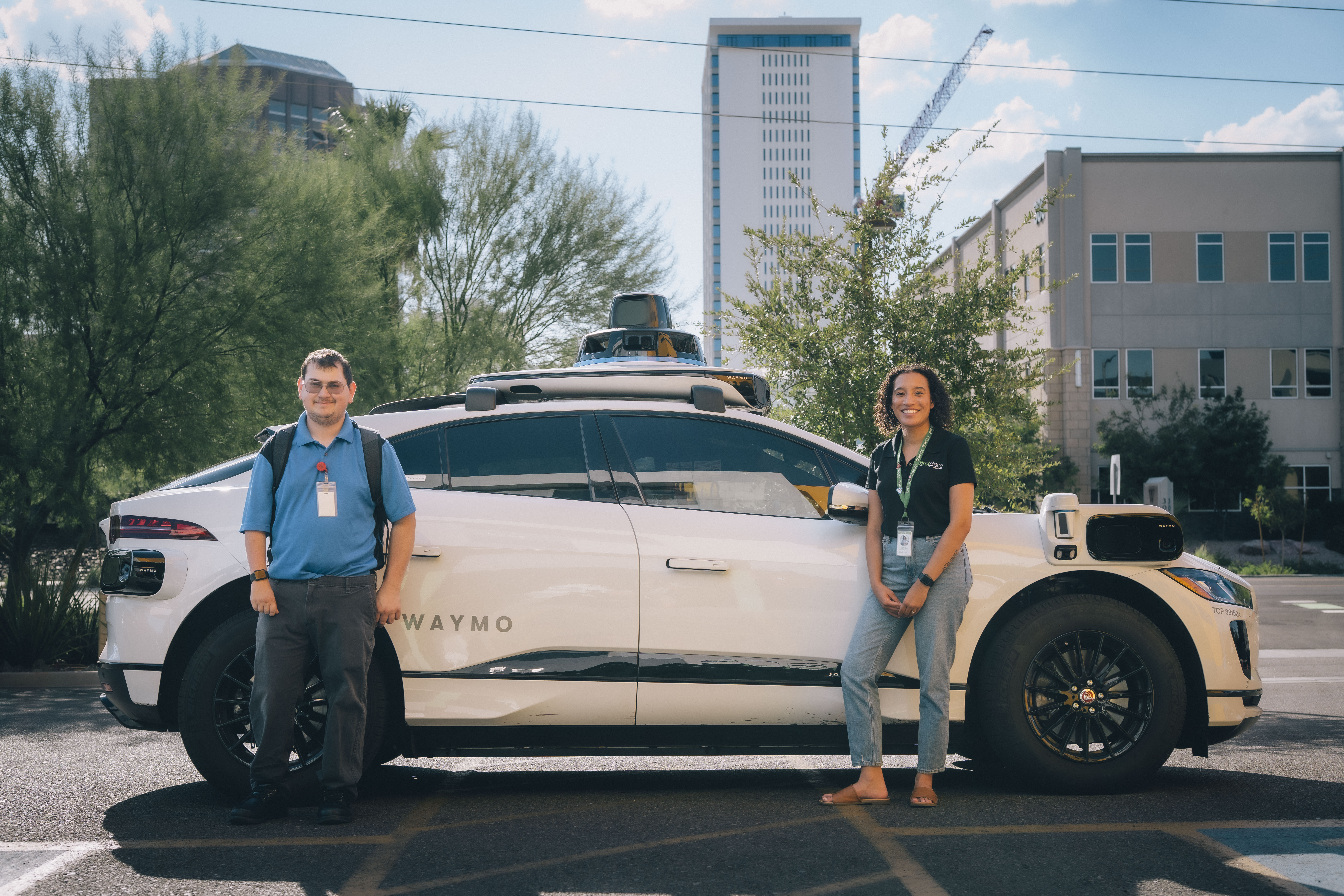 Eli and Natasha standing beside a Waymo autonomous vehicle, smiling toward the camera