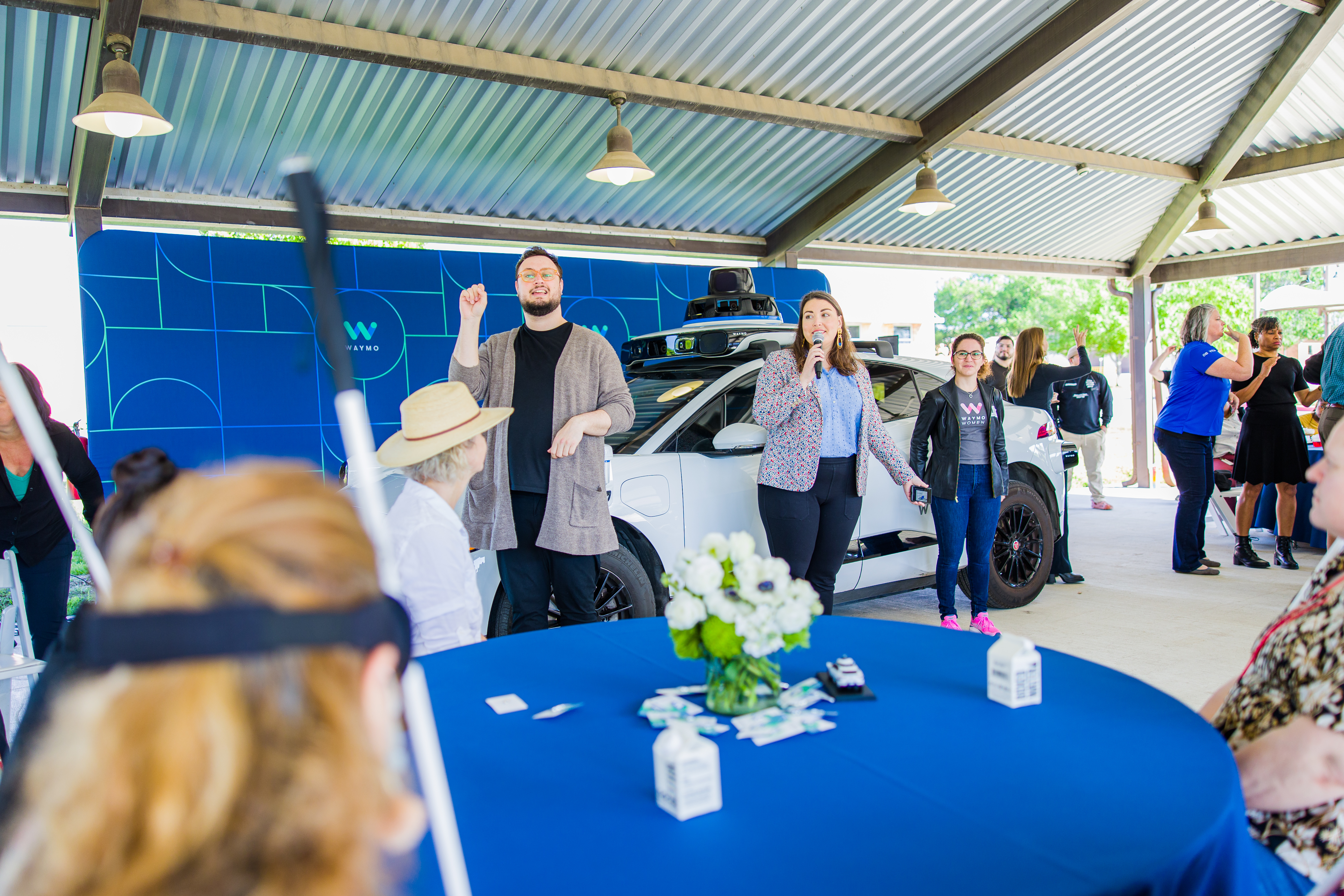 Representatives from Waymo standing next to an ASL interpreter in front of a crowd, next to a Waymo vehicle