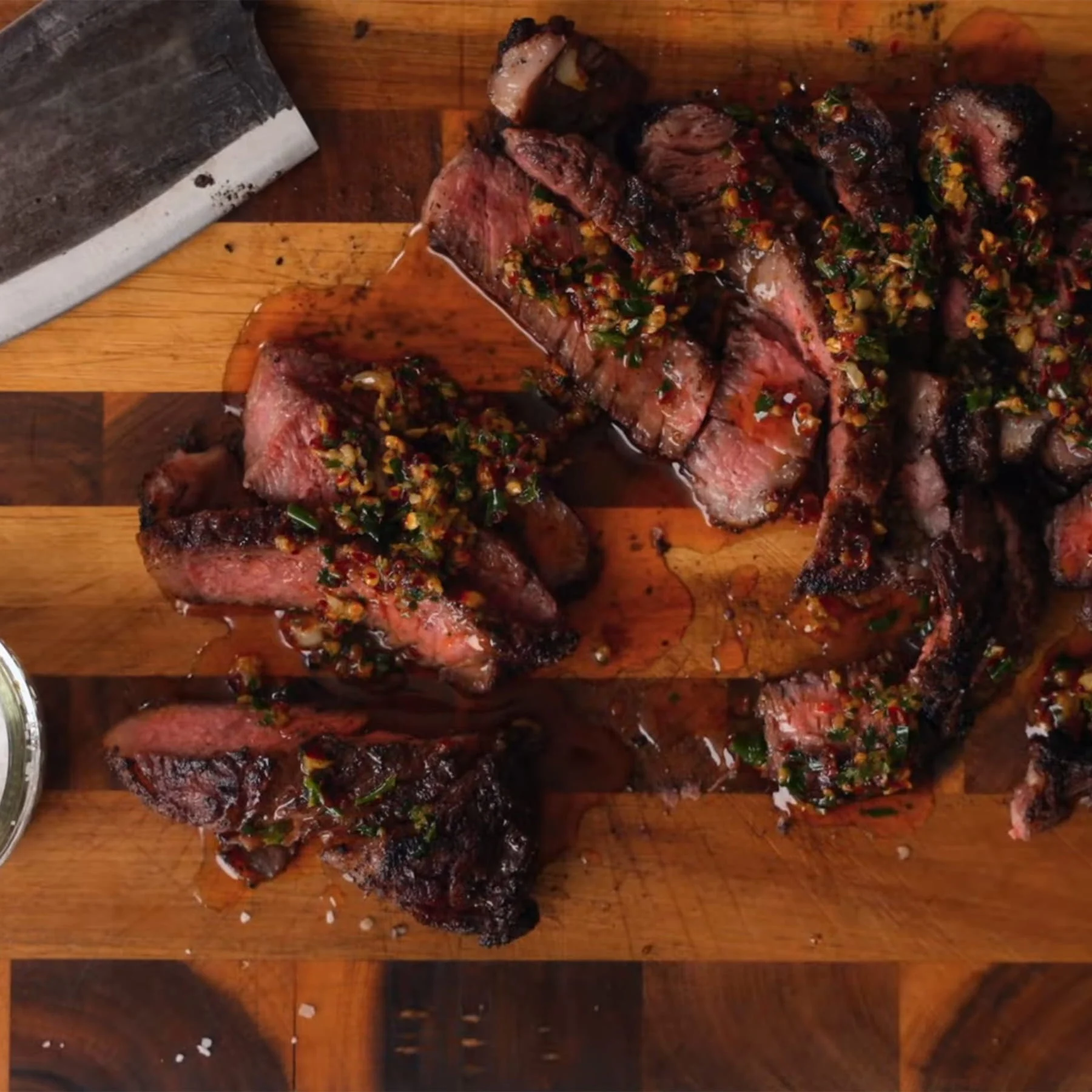 Slices of seared steak on a wooden cutting board with a knife and a ramekin of salt
