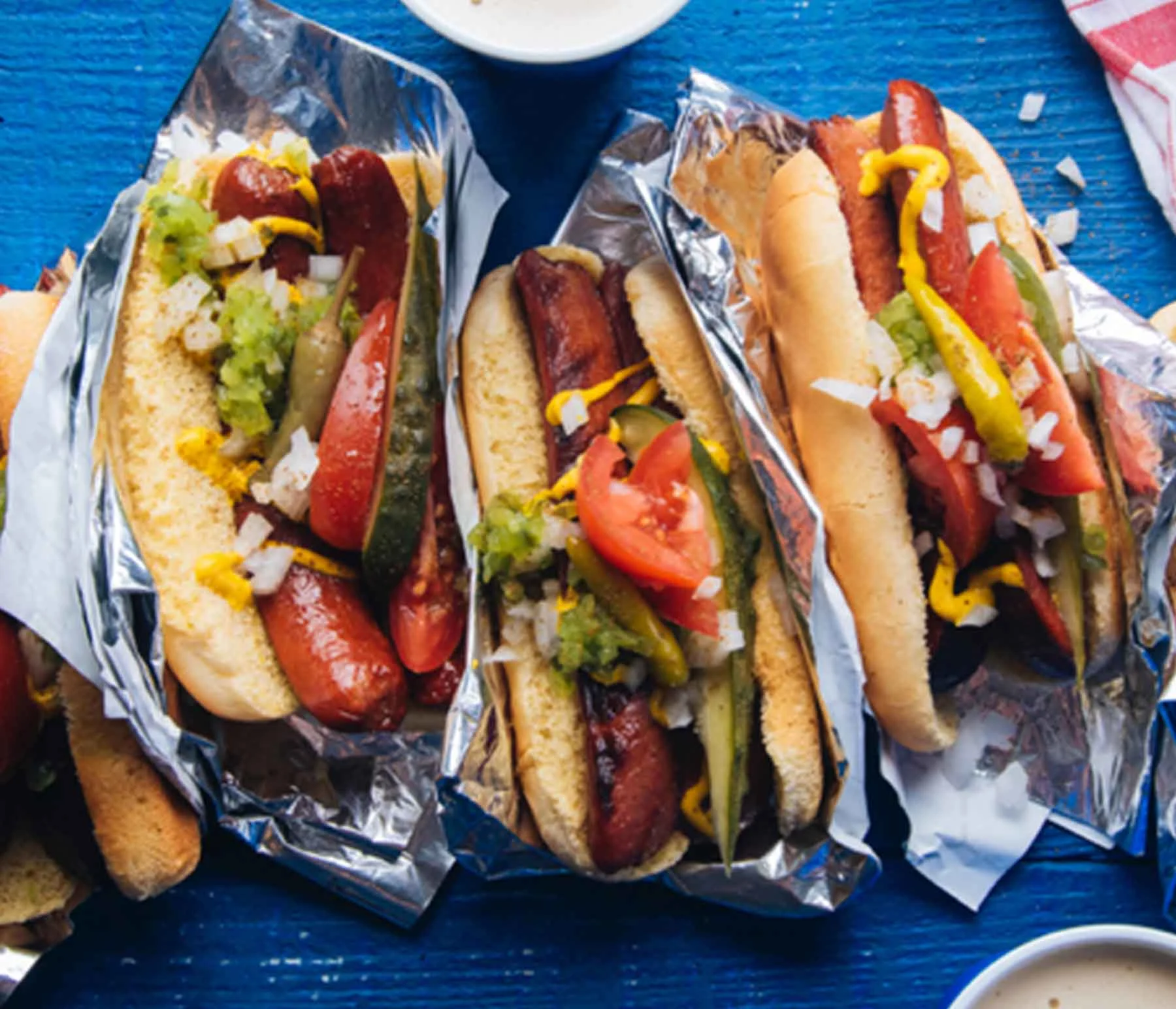Chicago-style hot dogs with tomatoes and relish, wrapped in aluminum foil on a bluw background