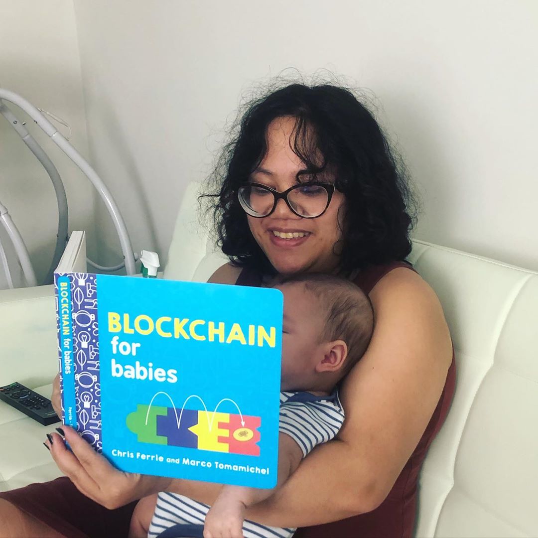 In this picture I'm reading a board book titled "Blockchain for Babies" to my child, who is dressed in a white-and-blue striped onesie. His face is obscured by the book.