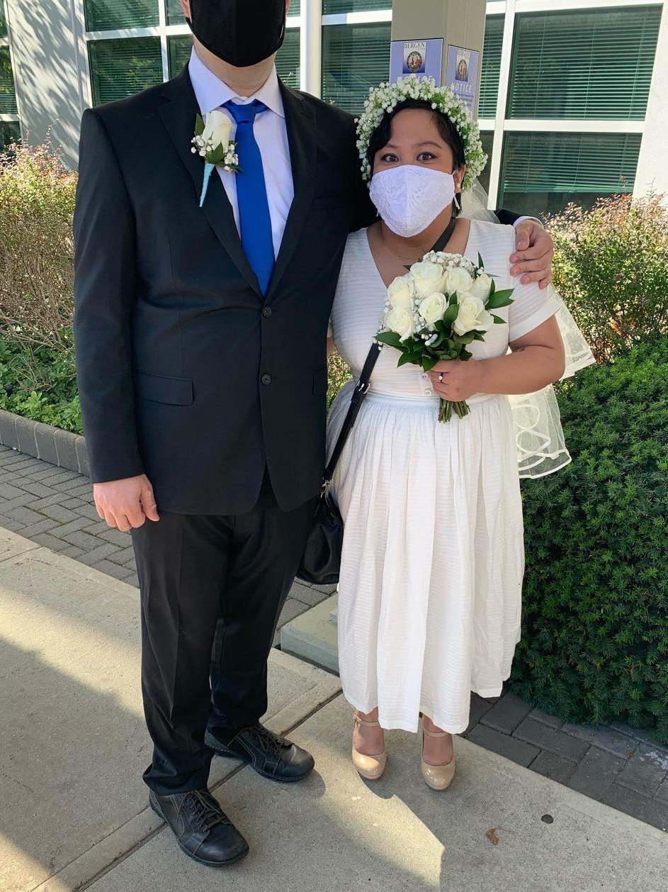 In this picture I'm with my spouse outside the county clerks office. I'm wearing a white dress, a white cloth face mask, a white veil, a crown of artificial baby's breath flowers, a black gold and moissanite engagement, ring and nude pumps. The pumps are quite a few shades lighter than my skin tone. I am holding a bouquet of white roses and a black purse. My spouse is in a black suit, a black cloth face mask, a white dress shirt, a royal blue tie, a white single rose boutonniere with baby blue ribbon, and some clean, but well-worn dress shoes. His eyes are out of frame, and his face mask obscures the rest of his face.