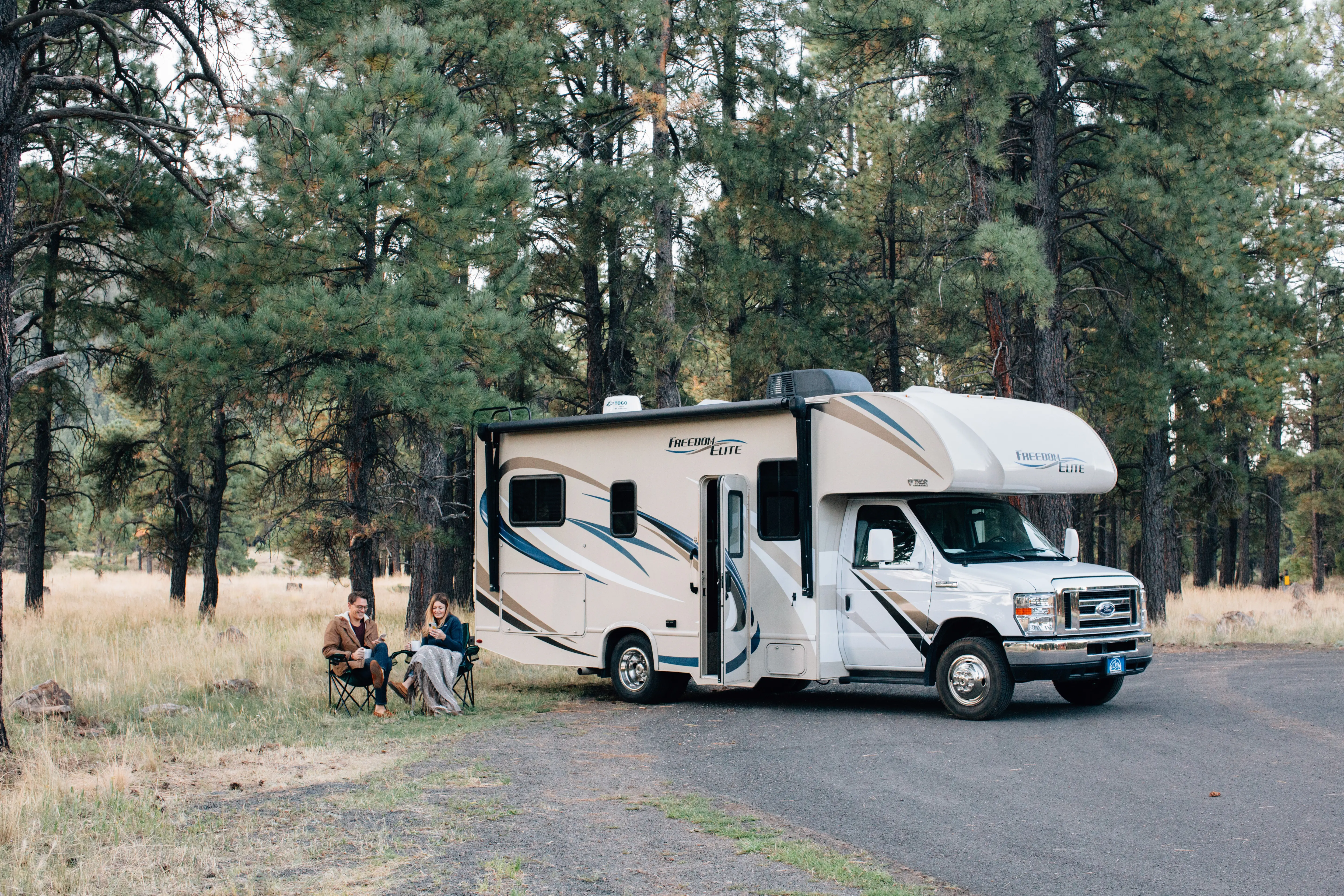 Two people sitting outside their RV with pine trees in the background.