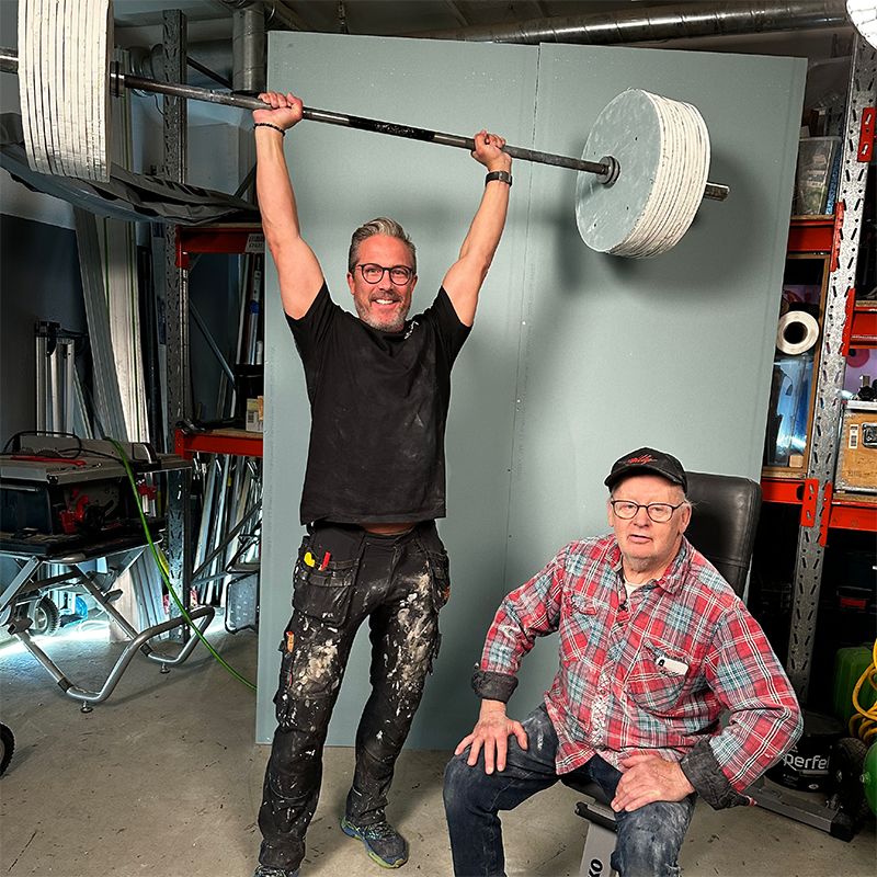 Verkstadsmiljö med svartklädd man som lyfter tung stång över huvudet, rutig skjortklädd man sitter.