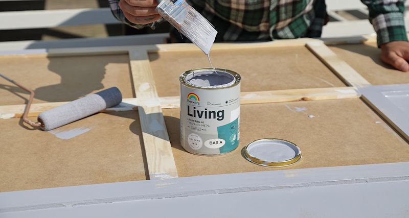 Hand med pensel droppar grå Beckers Living-färg på träram, roller bredvid.