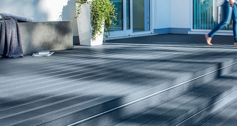 Mörkgrå terrass med komposittrall i solljus, skuggor, betongbänk, kruka och barfota ben