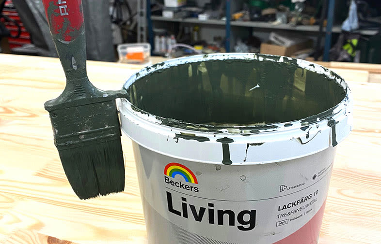 Burk med mörkgrön lackfärg "Beckers Living" och pensel med grön färg på träbord i verkstadsmiljö