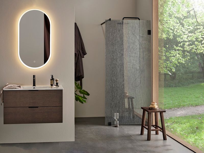 Modernt badrum med mörk träkommod, upplyst oval spegel, dusch bakom mönstrat glas och grön utsikt.