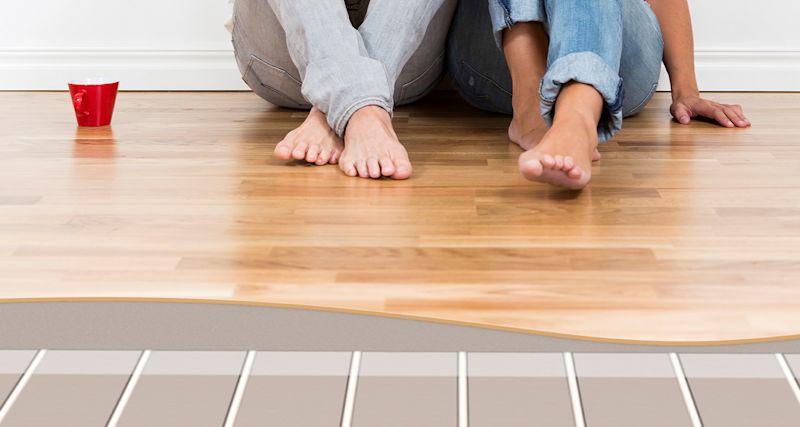 Två personer sitter barfota på ljust trägolv med röd mugg, underliggande golvvärmeskikt syns.