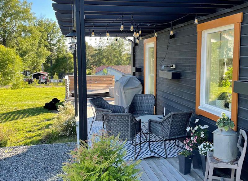 Altan med svarta korgstolar under pergola vid svart hus med orange fönster och trädgård med hund