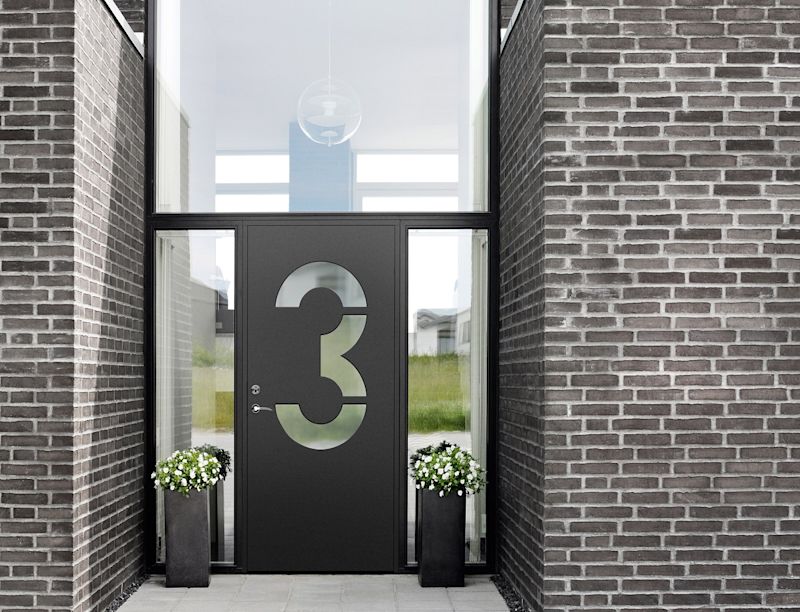 Svart ytterdörr med urfräst 3, smala glasfönster, tegelvägg och vita blomkrukor.