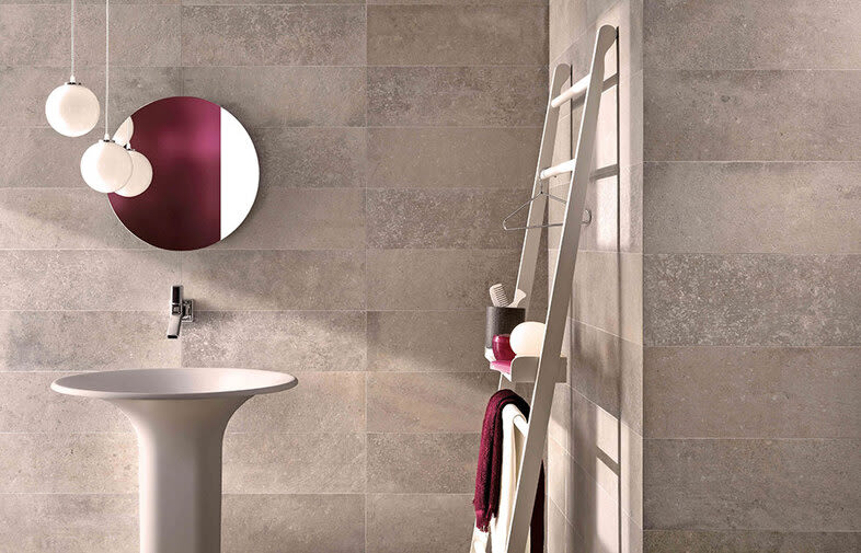 Modernt badrum med grå kakel, vitt handfat, rund spegel med vinröd halva och stege med handduk.