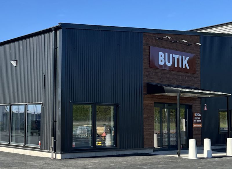 Svart korrugerad butikslokal med träpanel, röd skylt ”BUTIK”, glasdörrar och vita pollare.