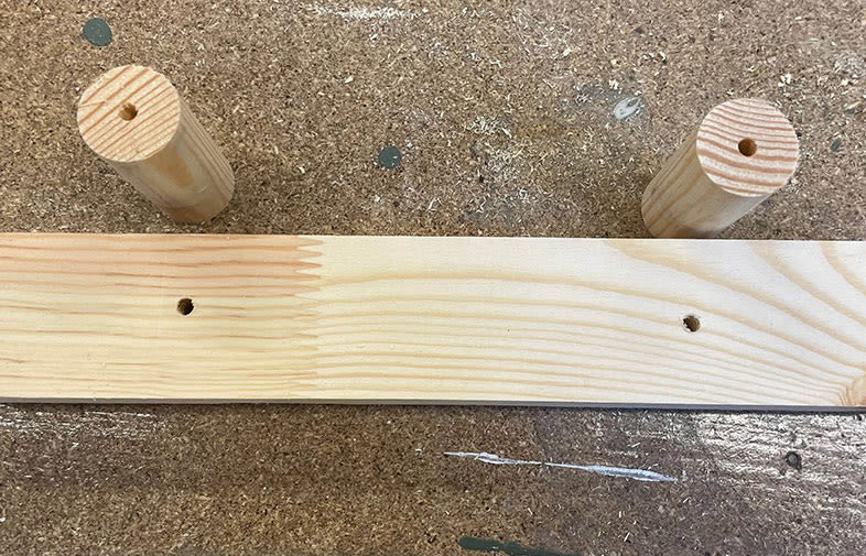 Ljust furubräda med två borrade hål och två träpluggar på dammig arbetsbänk.