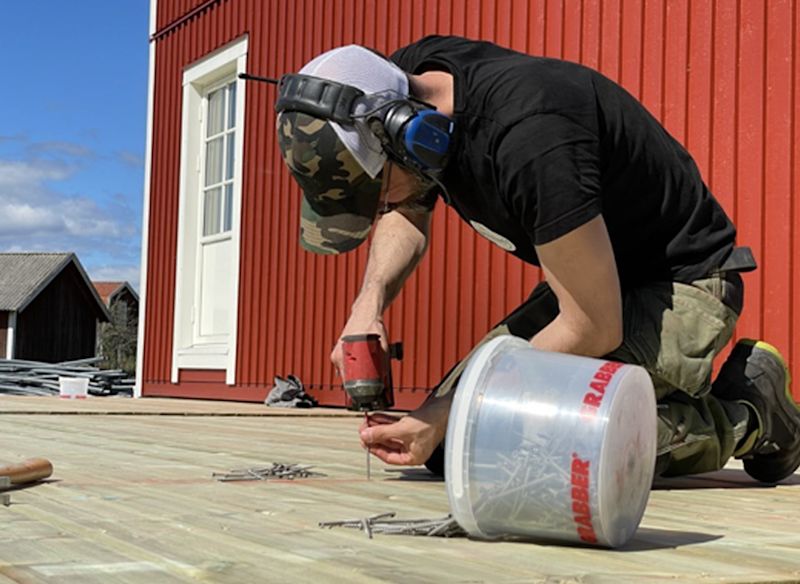 Snickare med hörselskydd skruvar trätrall framför rött hus med vit dörrkarm.