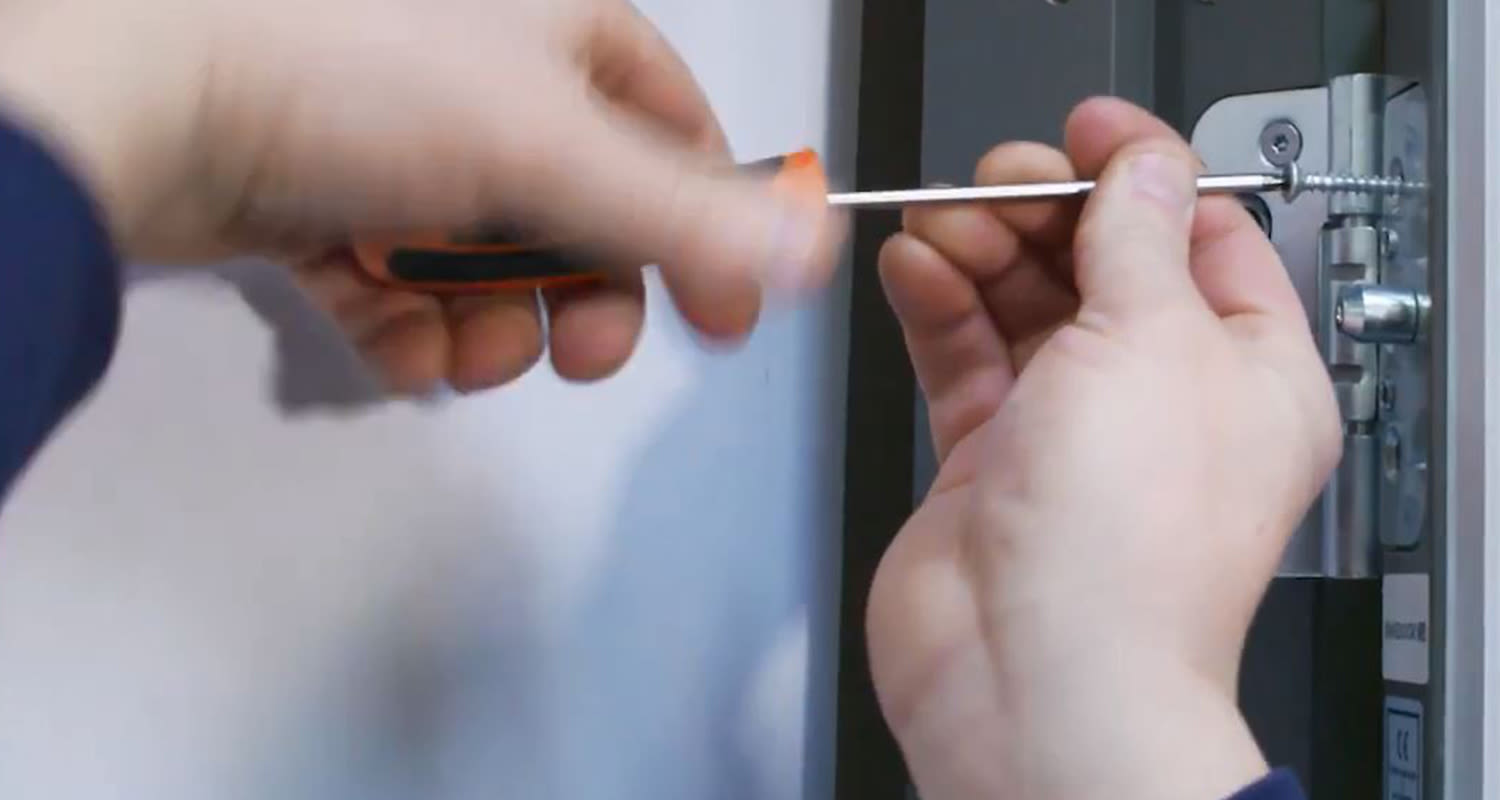 Närbild på händer som drar skruv i silverfärgat dörrgångjärn med orange-svart skruvmejsel