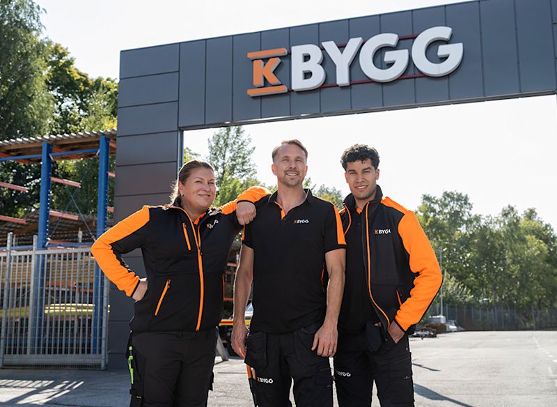 Tre byggarbetare i svartorange kläder poserar framför lagerhyllor fyllda med träplankor.