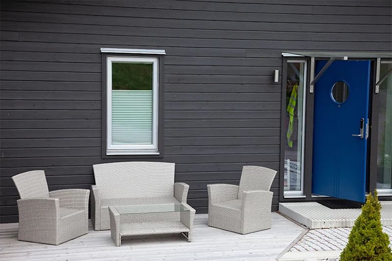 Mörkgrå träfasad med vitt rottingmöblemang på ljus altan framför blå dörr och fönster