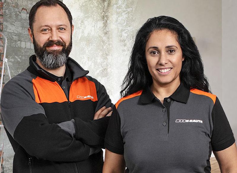 Två hantverkare i grå-orange K-Rauta-tröjor framför en rå vägg med stege.