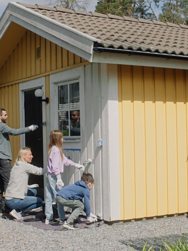 Familj målar vita brädor på en gul trästuga med roller och handskar.