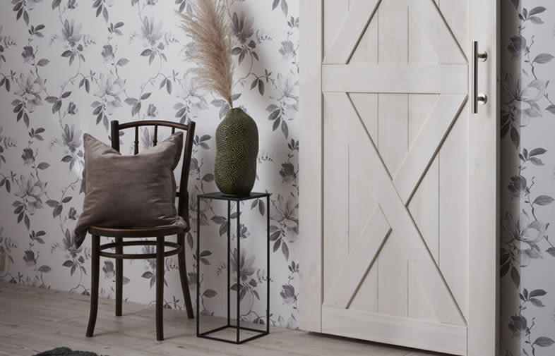 Brun trästol svart sidobord med grön vas och pampasgräs framför blommönstrad tapet och vit trädörr