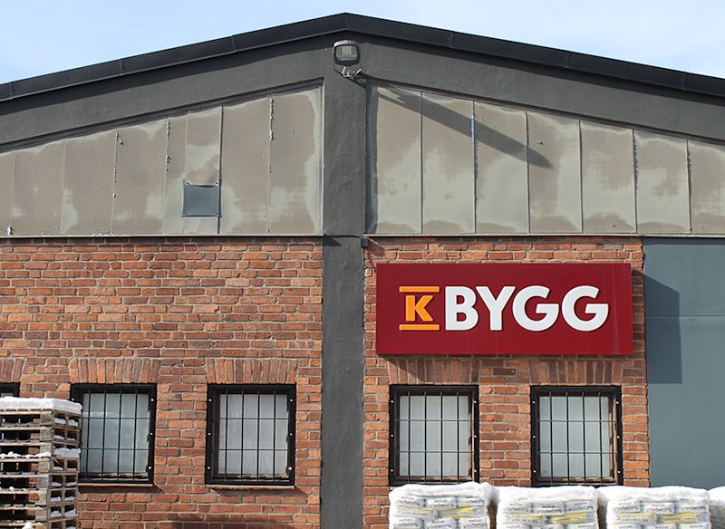 Röd KBYGG-skylt på tegelvägg med grå vindskiva, svarta gallerfönster och snötäckta säckar.