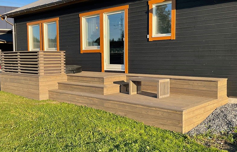 Modern träaltan med inbyggd bänk och småbord framför svart hus med orange fönsterfoder