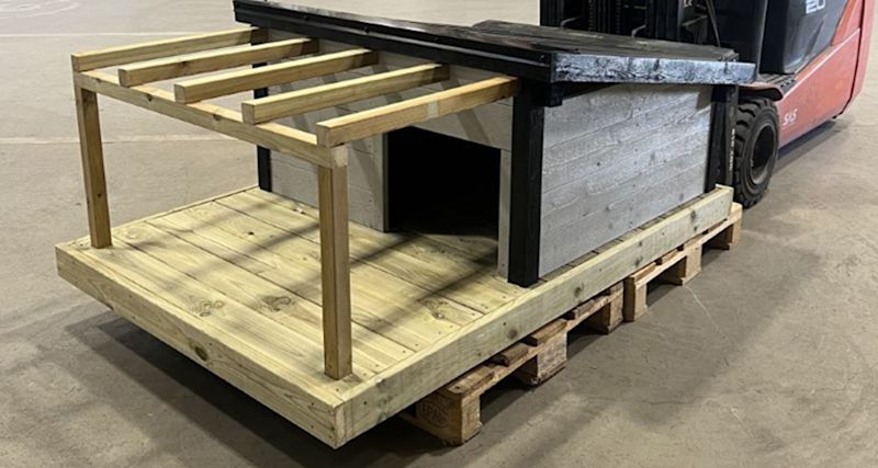 Ljus träplattform med överliggande reglar och grå låda på pall inomhus vid röd truck.