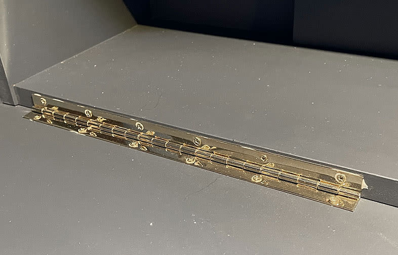 Långt gyllene pianogångjärn monterat med skruvar på svart träpanel.