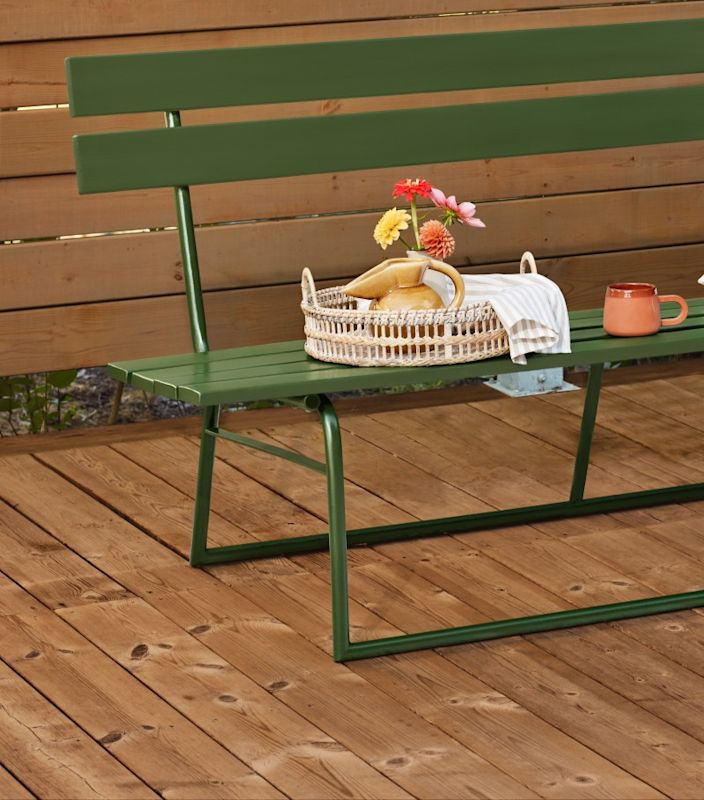 Grön bänk på trädäck med rottingbricka, kanna, randig duk, färgglada blommor och kopp