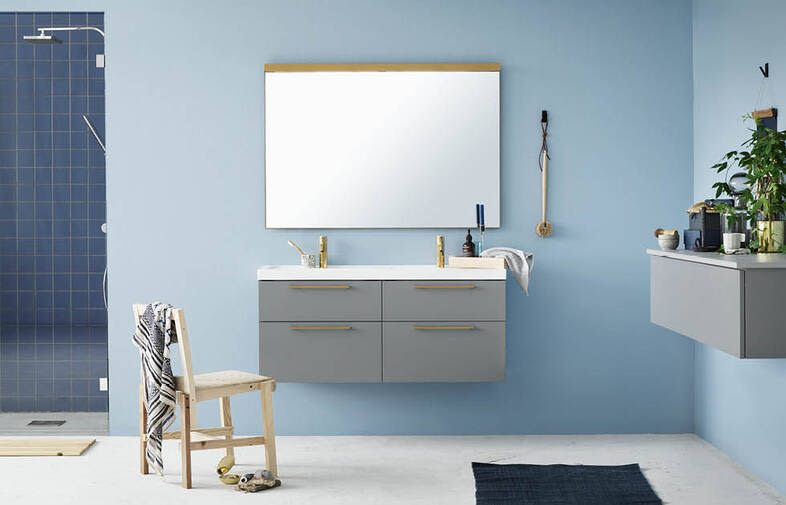 Badrum med ljusblå vägg, grå vägghängd kommod, guldfärgade kranar, stor spegel och blå kaklad dusch.