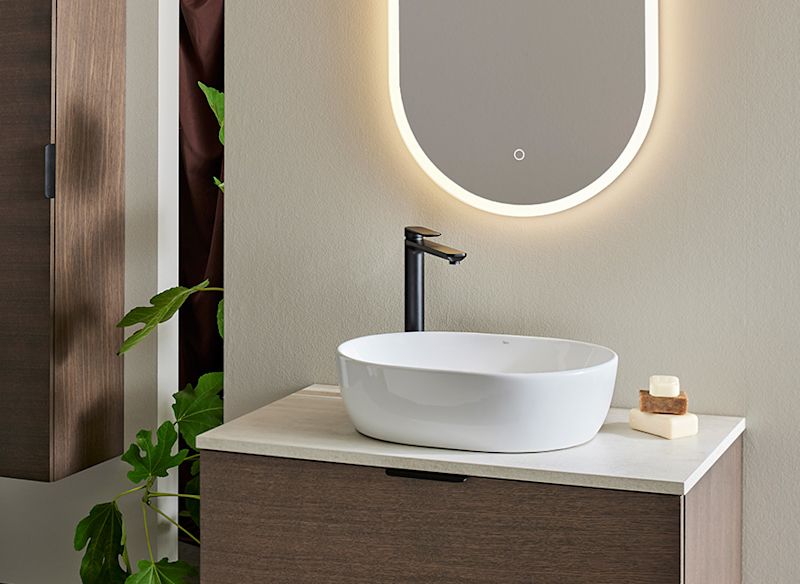 Vit oval handfat med svart kran på träskåp, upplyst oval spegel, grön växt och beige vägg.