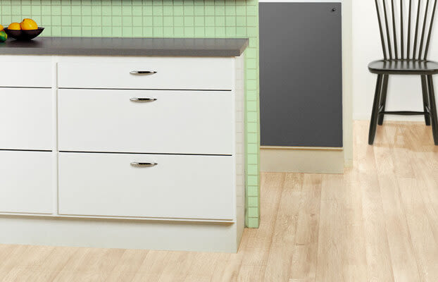 Vitt köksskåp med grå bänkskiva, grön kakelvägg, fruktskål, svart stol och ljust trägolv.