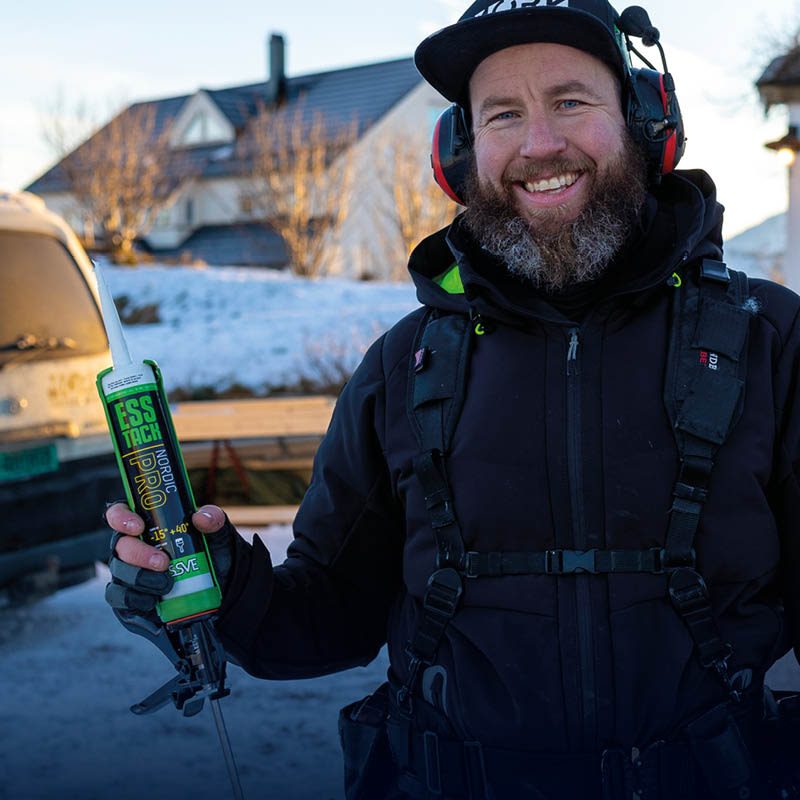 Man i svart vinterutrustning med hörselskydd håller grön limpatron i snötäckt byggarbetsplats.