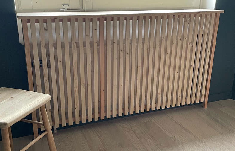 Ribbad radiatorskydd i naturträ framför vitt fönster, mörkgrön vägg, ljust trägolv och träpall.