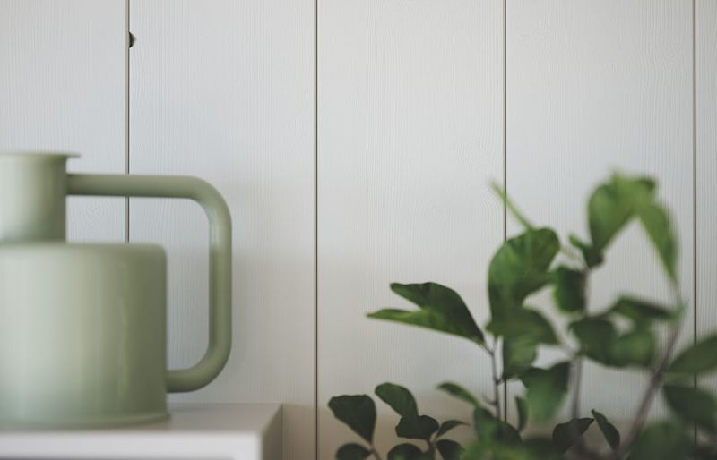 Pastellgrön vattenkanna på vit hylla mot vit trävägg, suddig grön krukväxt i förgrunden.
