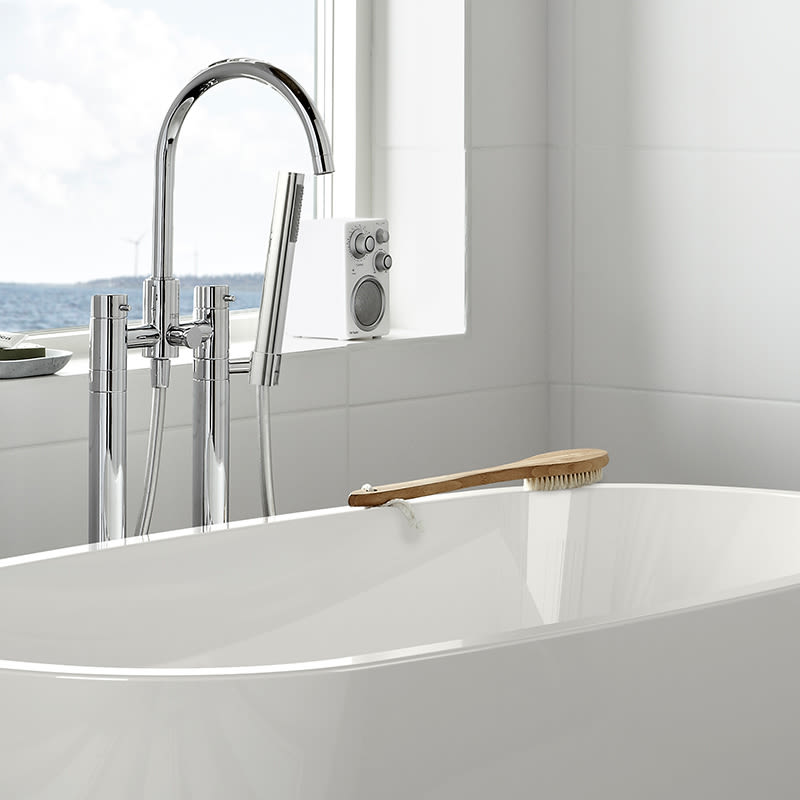 Vit fristående badkar med kromblandare och träborste, fönster med havsutsikt och vindkraftverk.