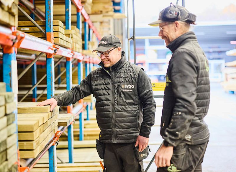 Två lagerarbetare i svarta västar vid blå-röda hyllor med träbrädor i byggvaruhus