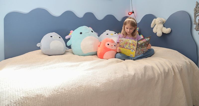 Flicka läser bok på blå säng med ljus täcke och fyra färgglada runda gosedjur uppradade