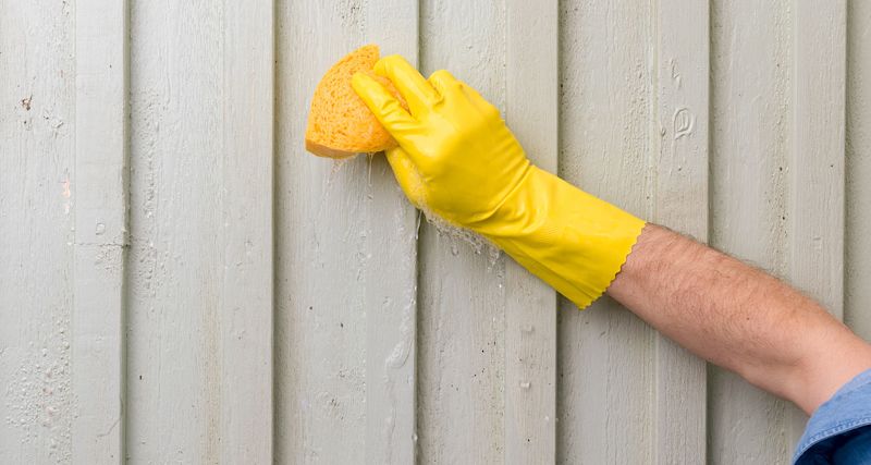 Hand i gul gummihandske torkar grå träpanel med gul svamp, vattendroppar