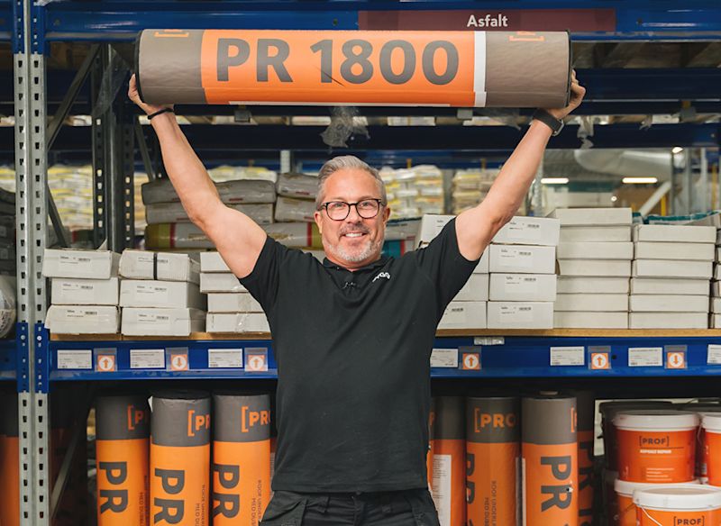 Person som håller upp en stor rulle med texten “PR 1800” tryckt på den. Rullen är orange och brun, och verkar vara ett byggmaterial, troligen en underlagsduk eller takpapp. I bakgrunden syns hyllor fyllda med fler rullar av samma typ samt staplade kartonger.