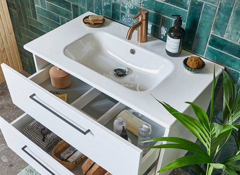 Vit bänkskiva med handfat, mässingskran, öppna lådor, grön kakelvägg och växt.